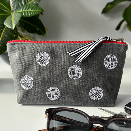 Gray pouch with white spherical patterns and a red zipper on a surface with sunglasses and a phone.